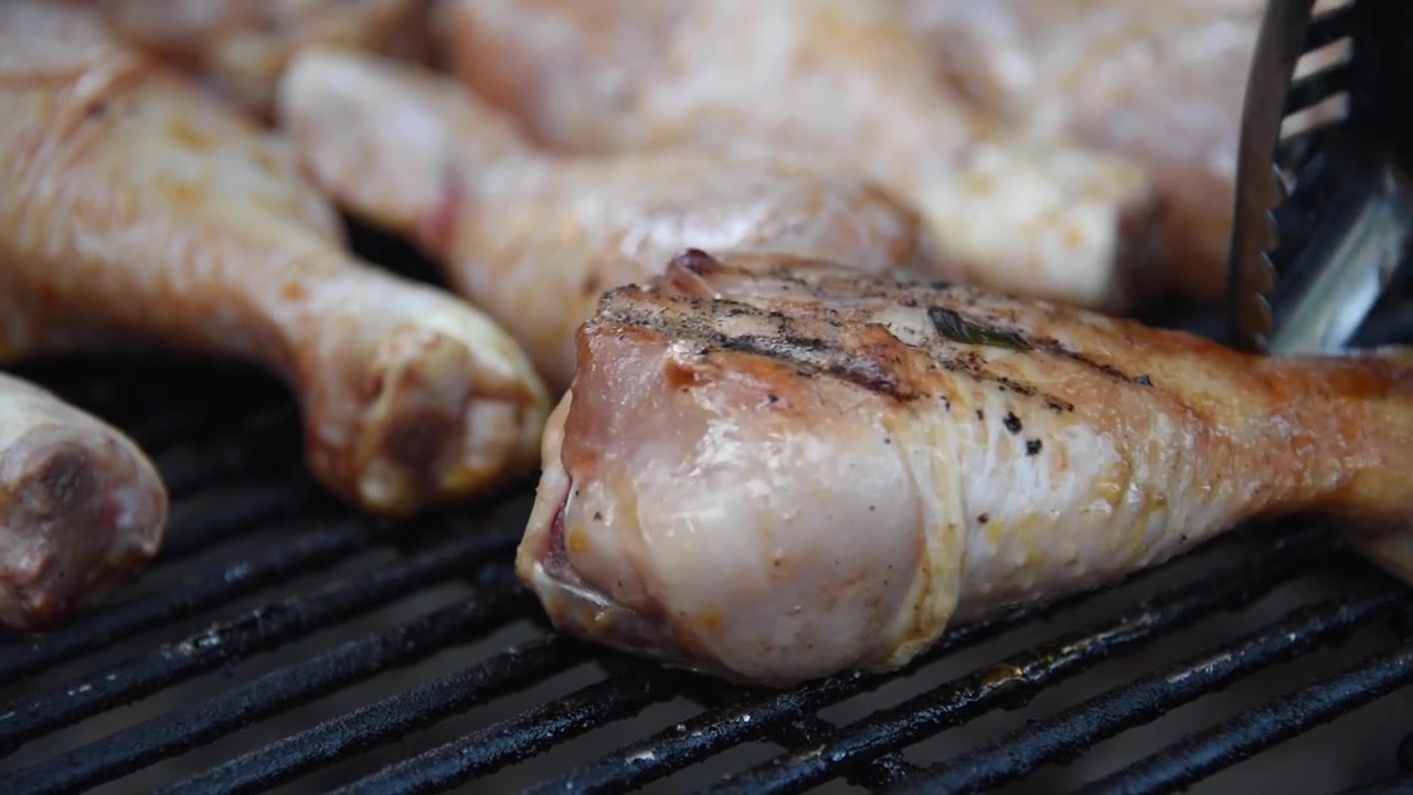Primo piano di fusi di pollo marinati che cuociono su un barbecue. Una spatola di metallo gira la carne mentre sfrigola sulla griglia calda dell'affumicatore.
