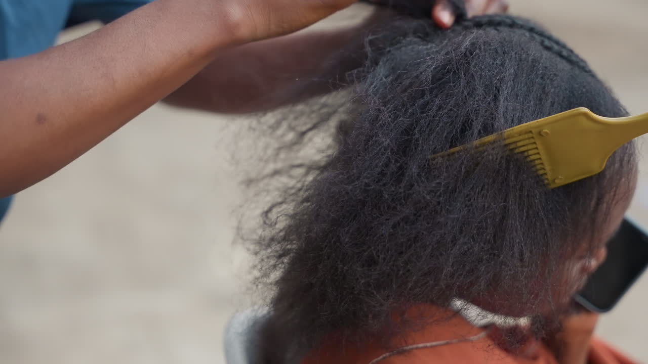 Close-up pengasuh berkulit hitam menata rambut bertekstur dengan sisir garpu kuning, gerakan mengembang dan menghaluskan yang lembut, suasana penuh perhatian, nuansa salon rumahan, rutinitas keluarga yang menenangkan, dan detail perawatan rambut.