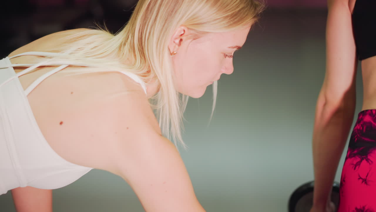 blond kvinde og partner løfter vægte med spotterstøtte, lyserøde leggings og sort croptop, dynamiske dødløft-gentagelser på fitnessgulvet, fokuserede ansigter, kontrolleret nedstigning, udstyr synligt