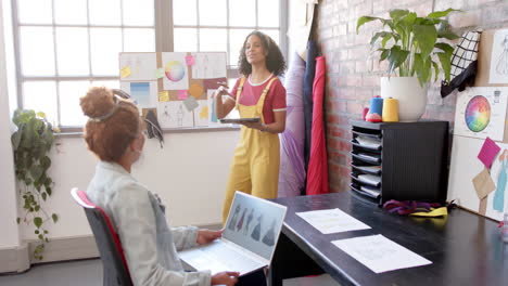 Diverse-female-fashion-designers-using-laptop-and-tablet-discussing-designs-at-studio,-slow-motion