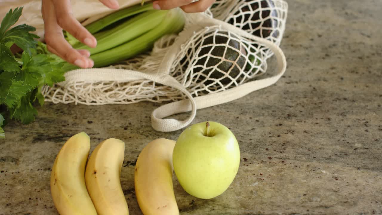 Mãos e antebraços alcançando uma sacola de compras de malha e criando uma exibição organizada de bananas e maçãs no balcão
