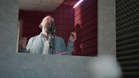 ein mann singt leidenschaftlich in einem aufnahmestudio, trägt schwarze kopfhörer und ein blaues hemd mit langen ärmeln. er spielt mit geschlossenen augen und handbewegungen, umgeben von roten akustischen platten