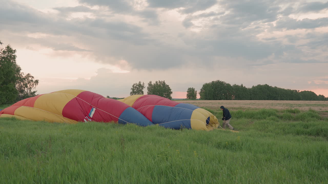 person in black sweater and gloves pulling deflated hot air balloon envelope across grassy field at sunset sky with distant tree line and vehicles during preflight pack preparing flight materials
