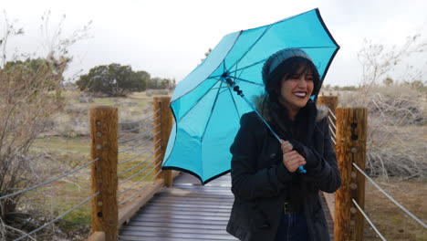 Una-Mujer-Bonita-Caminando-Bajo-La-Lluvia-Y-El-Mal-Tiempo-Ventoso-Durante-Una-Tormenta-Con-Un-Paraguas-Azul-Mientras-Sonríe-Y-Se-Ríe