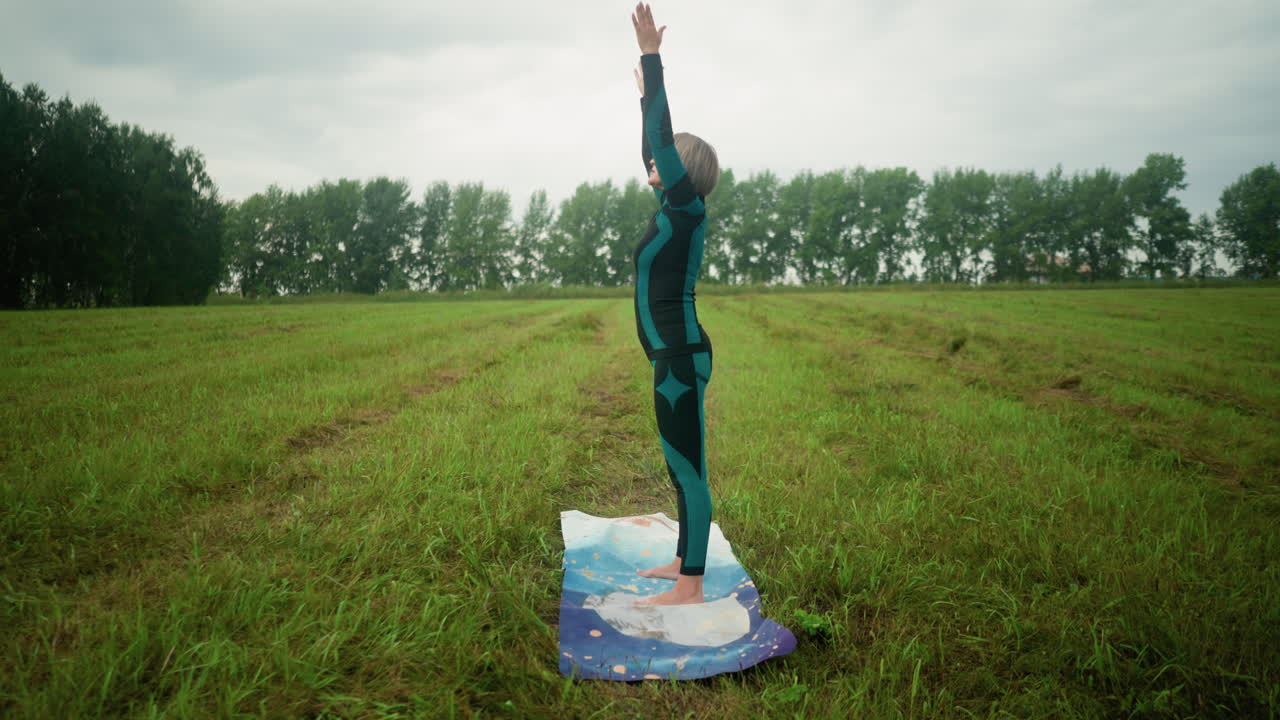 야외에서 다채로운 매트 위에 서있는 여자의 측면 뷰, 머리를 약간 들어 올리고, 손을 천천히 펴고, 구름 하늘 아래에서 요가를 연습하는 동안 얼굴로 내려 올립니다.