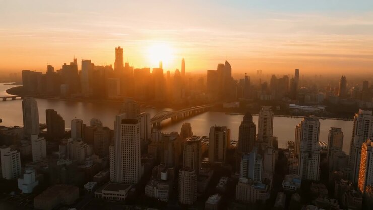 Movimento aéreo lento atravessa torres sombreadas em direção ao rio e à ponte, enquanto a luz solar quente silhueta torres altas distantes e reflete suavemente na água calma sob um céu pálido