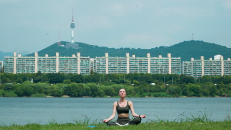 Slender-young-woman-sitting-on-yoga-mat-in-lotus-position-and-raising-hands-up-outside-in-Han-river-city-park