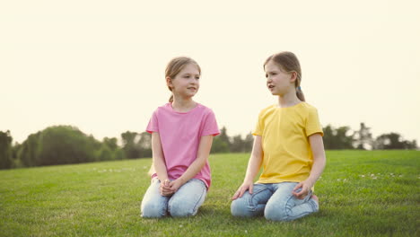 two pretty little sisters talking together, looking around and pointing something while kneeling on grass at park