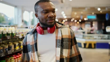 A-happy-and-confident-man-with-Black-brunette-skin-color-in-a-checkered-shirt-and-red-wireless-headphones-walks-along-the-counter-in-a-modern-spacious-and-sunny-supermarket-and-looks-at-the-goods-on-display