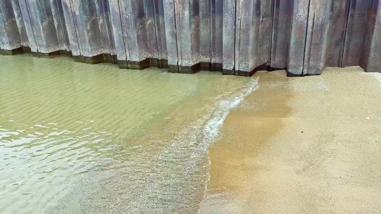 Water meets sand near a wall during low tide at a coastal area in mid-afternoon light