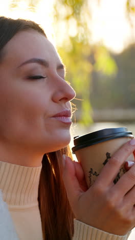 enjoying hot coffee from a paper cup near the river. long-haired attractive lady sitting under the tree in the rays of bright autumn sun. blurred backdrop. vertical video