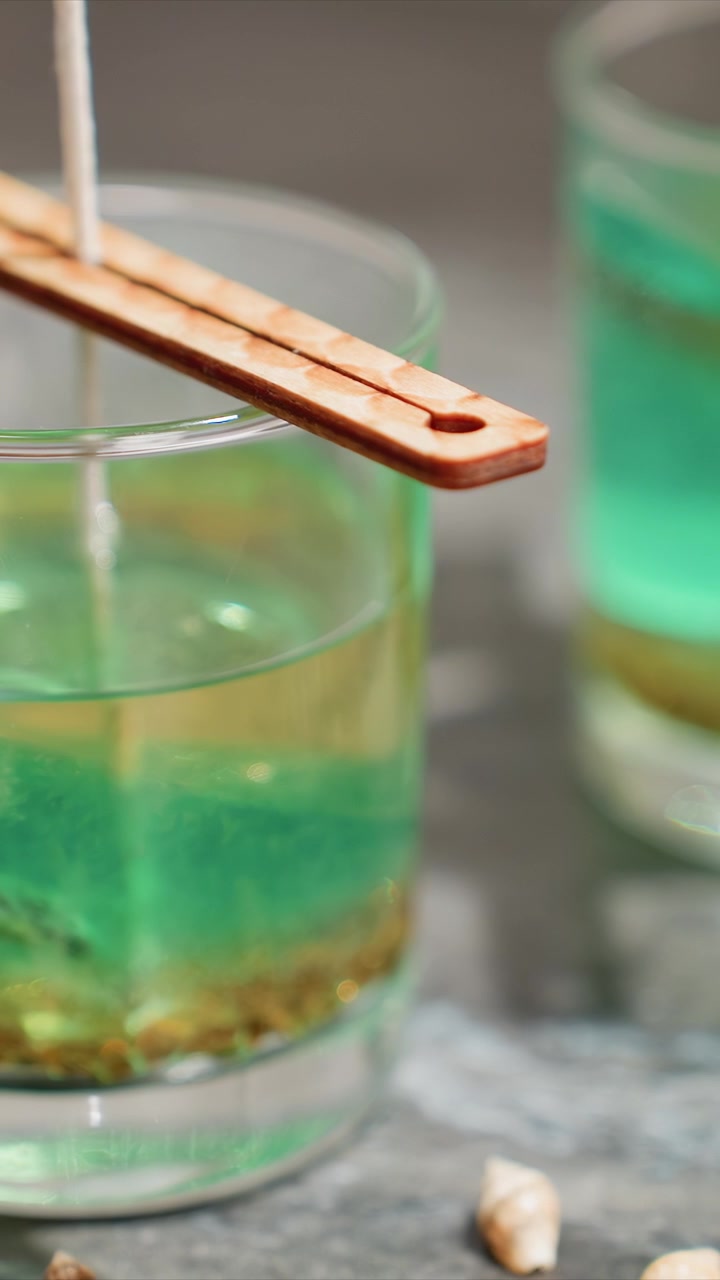 Primo piano di cera di candela verde versata in un contenitore di vetro con stoppino e stampo di legno visibili. La cera scorre costantemente mostrando dettagli di texture come granuli sul fondo mentre un altro simile.