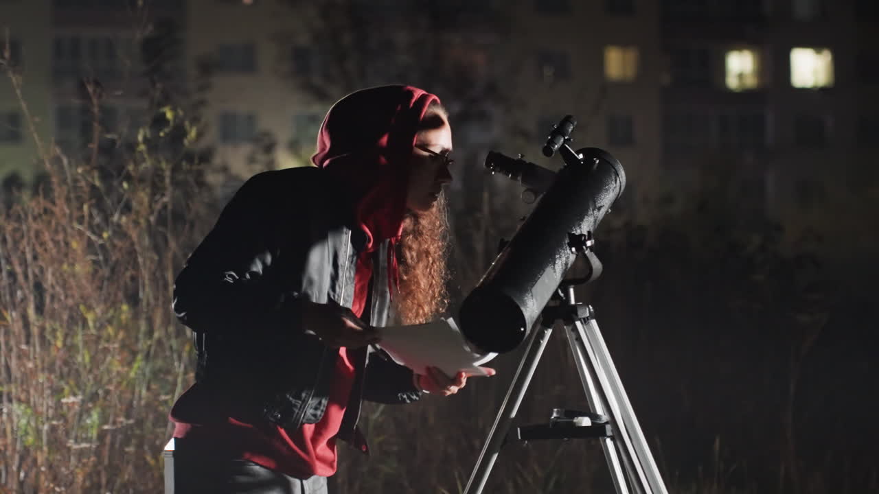 Astronomo bianco con la barba calibra il telescopio di notte, modificando cercatore e treppiede, consulta una piccola carta stellare e appunti, felpa rossa con cappuccio e lunga barba illuminate dalle finestre distanti di un appartamento, riflettore