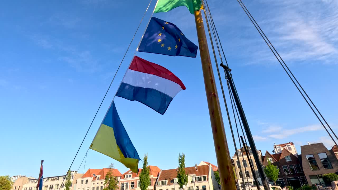Multiple flags wave on a boat mast under clear blue sky, historic Dutch buildings behind