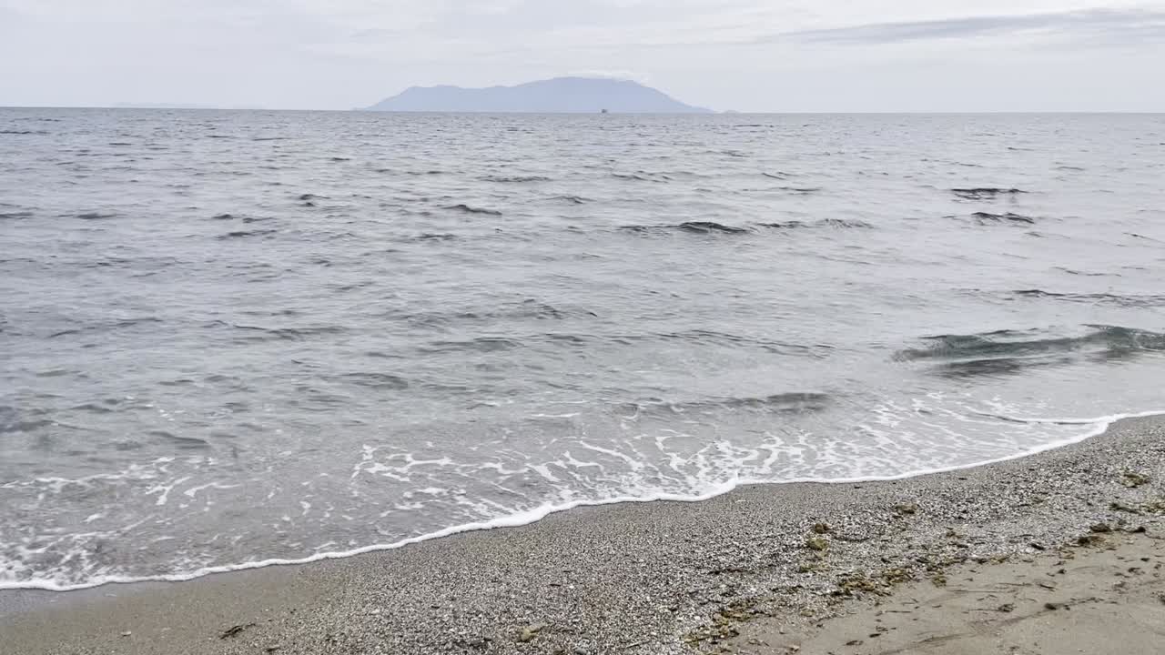 Peaceful seaside view of small waves washing onto a sandy beach with a distant island on the horizon, ideal for travel, relaxation, and nature themes