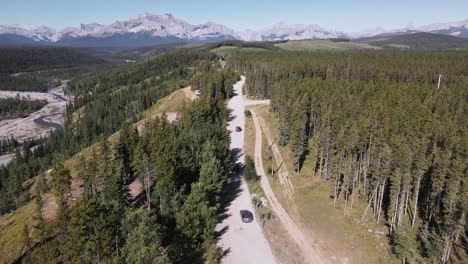 multiple cars driving along a dusty logging road through a boreal forest with a majestic rocky mountain backdrop