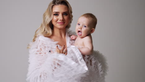 attractive blonde long-haired woman with a naked child in hands. loving mom kisses and touches her baby tenderly. white backdrop.