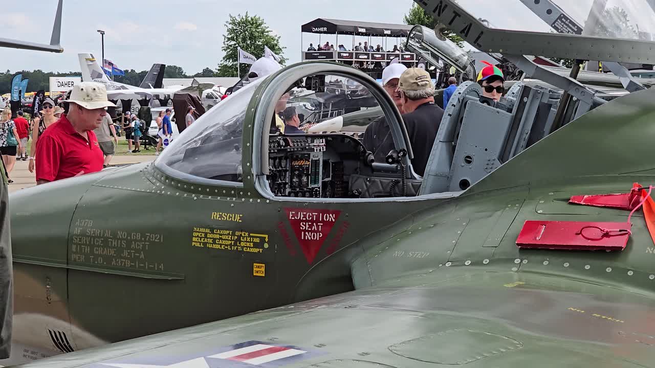 Close-up of a vintage military aircraft cockpit with ejection seat warning labels, displayed at EAA AirVenture in Oshkosh, WI. Attendees observe in the background.