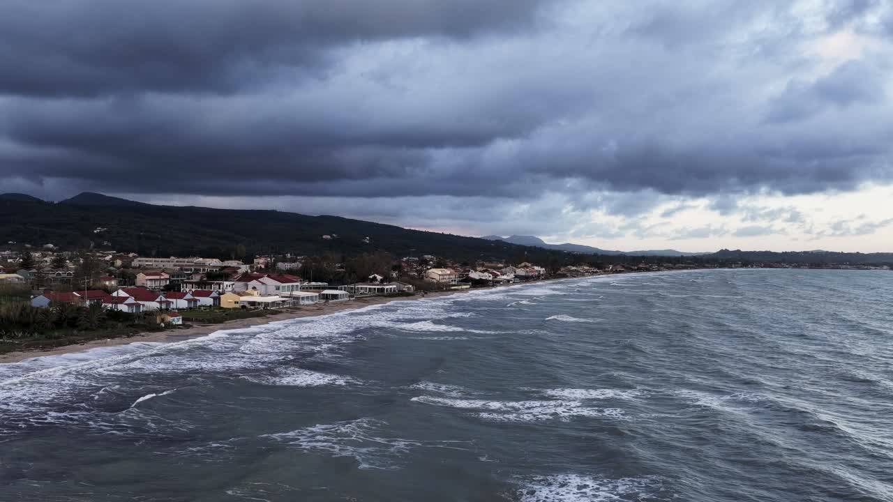 Vista aerea di onde tempestose che si infrangono sulla spiaggia di Acharavi a Corfù Nord, Grecia, sotto nuvole scure e drammatiche, mostrando la costa frastagliata e l'architettura dei villaggi costieri durante l'inverno.