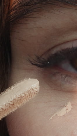 woman applies pastel concealer on soft skin under eye with applicator. young female person covers dark circles under eyes before makeup macro