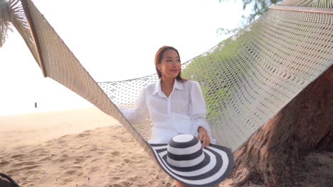 a young pretty woman rocks gently in a rope hammock slung over a sandy southeast asian beach