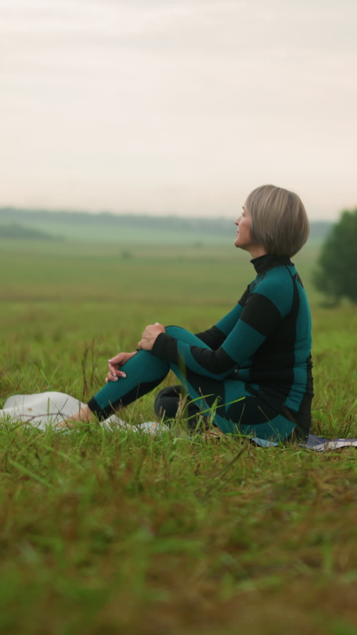 안개 잔디에 있는 요가 매트 위에 앉아 무을 들고 멀리 쳐다보며, 자연에 둘러싸여 평온함을 받아들이며, 평화로운 마음김 명상에 종사하는 여성.