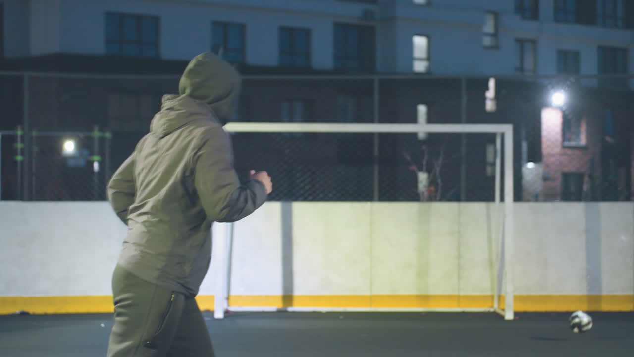 Player kicks ball with precision into goal post during nighttime soccer training session on outdoor field, ball rolls out after scoring under stadium lights, surrounded by urban buildings