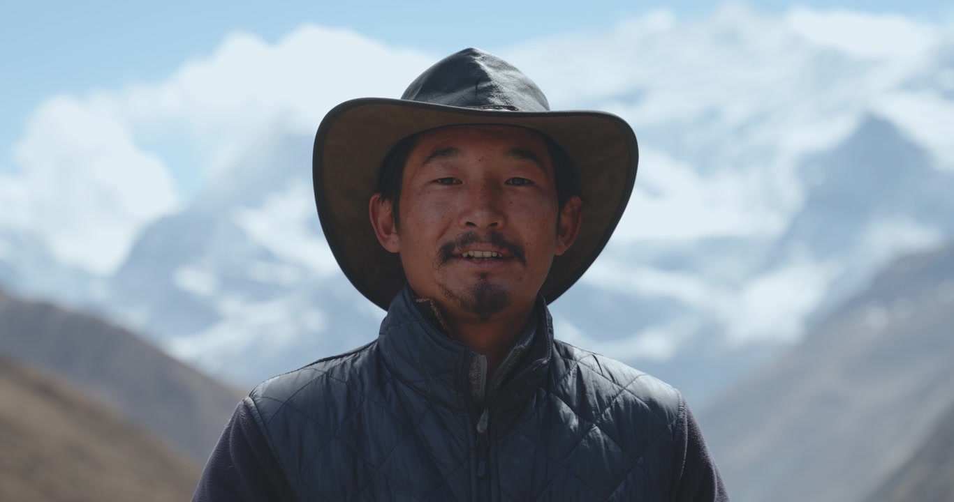 Annapurna CIrcuit, Manang Nepal, a smiling local Sherpa man wearing a cowboy hat and dark jacket, standing against the blurred background of the snow-capped Annapurna peak