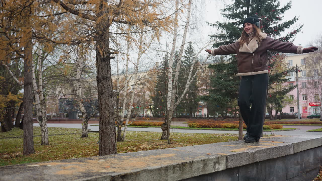 Confident lady wearing black knit cap brown shearling jacket black trousers balancing with arms outstretched while walking on wide stone surface in autumn park surrounded by bare trees and golden leaves