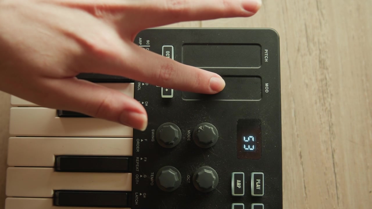 Primo piano dall'alto che mostra la sezione sinistra di una tastiera elettronica compatta mentre una mano preme e fa scorrere la striscia tattile accanto alle manopole rotanti, creando cambiamenti visibili di intonazione e modulazione nella configurazione musicale.