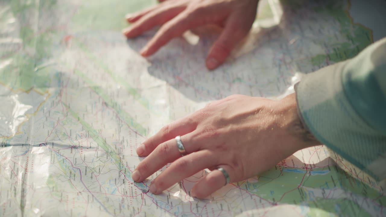 Hender på kart som planlegger rute, nærbilde av hender med ringer som sporer topografiske linjer, mykt sollys på sammenbrettet papirkart spredt på bakken, stille forventning under utendørs reiseforberedelse