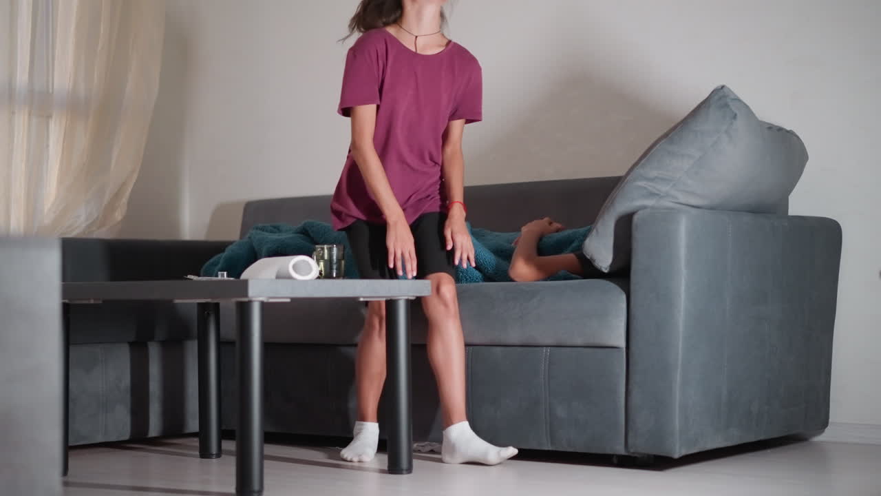 Niña pequeña con calcetines blancos sentada junto a su hermano dormido en el sofá, lo tapa suavemente con una manta, se levanta para irse mientras la medicación y el agua permanecen sobre la mesa; momento de cariño, amor fraternal.