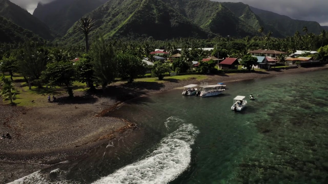 Un drone aereo segue una piccola imbarcazione che attraversa un canale di barriera corallina vicino a Teahupoo, Tahiti, con la sua scia che taglia una linea bianca attraverso le calme acque della laguna.