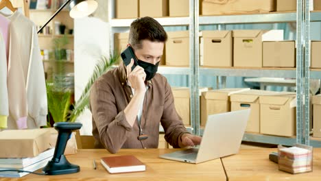 hombre diseñador caucásico con máscara negra sentado en la mesa y hablando en un teléfono inteligente mientras navega en línea en una computadora portátil