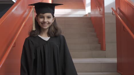 戴着帽子和长袍的快乐的幼儿园女学生的肖像,走下楼梯,拿着毕业证书,看着相机