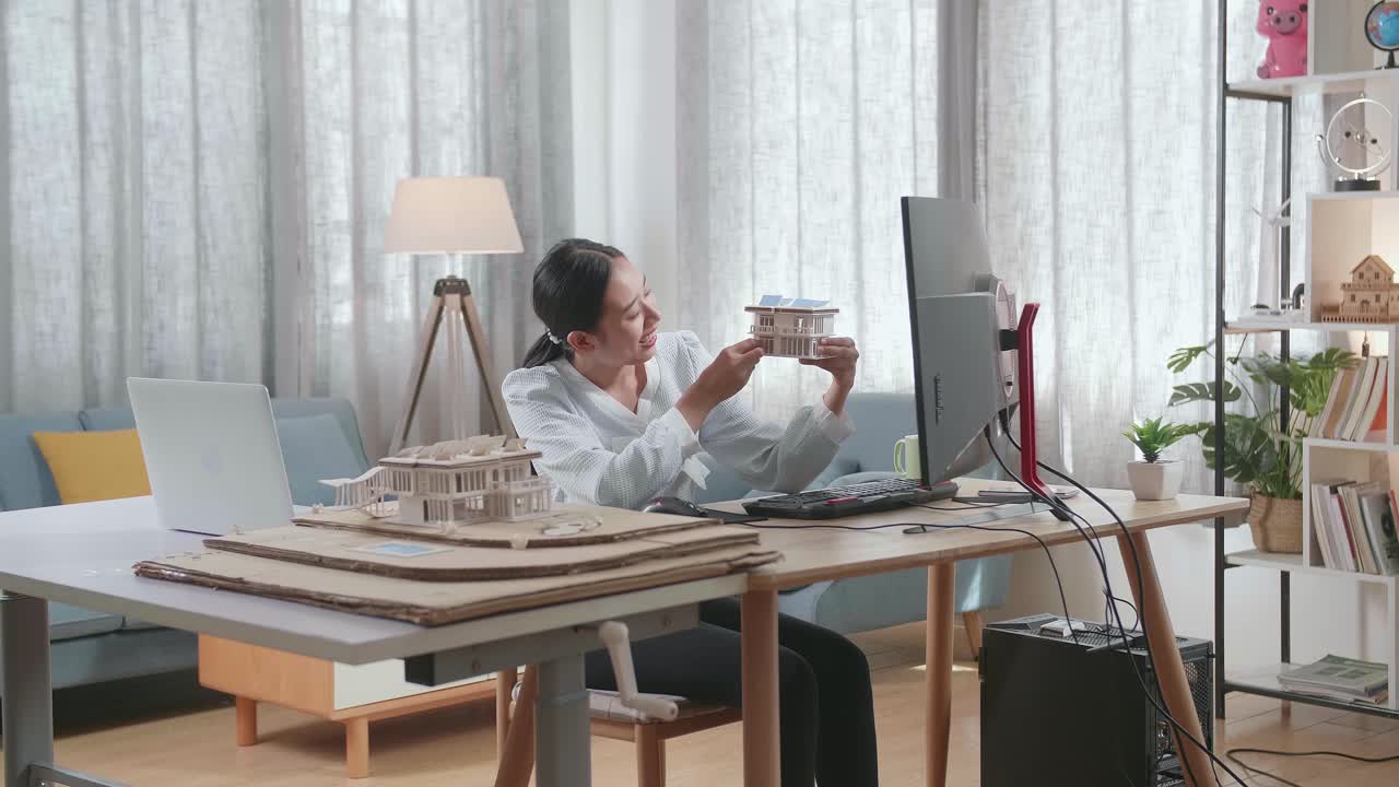 아시아 여성 엔지니어가 집에 있는 데스크에 있는 사진과 비교하기 전에 태양광 패널을 가진 작은 집 모델을 들고 보고 있습니다.