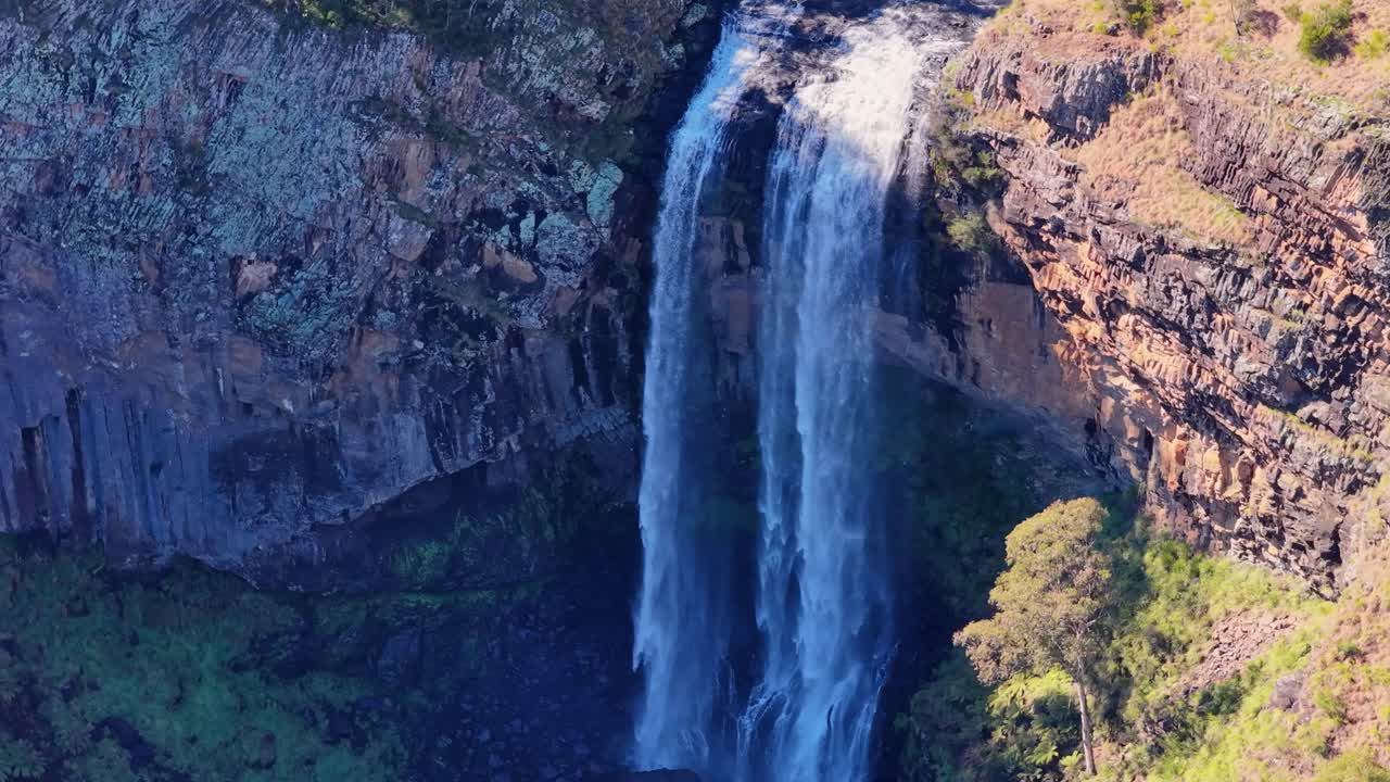 Drone footage captures Ebor Waterfall cascading over rocky cliffs into a lush green gorge under bright daylight, highlighting natural textures and dramatic scenery