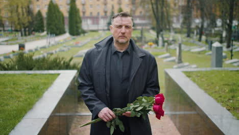 vista frontal del hombre con impermeable negro y traje sosteniendo rosas mientras camina en un cementerio