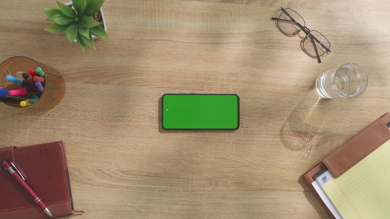 vista de arriba hacia abajo de una computadora portátil con pantalla verde simulada pantalla chromakey en un escritorio de oficina de madera al lado de la libreta con bolígrafos, gafas y un vaso de agua.