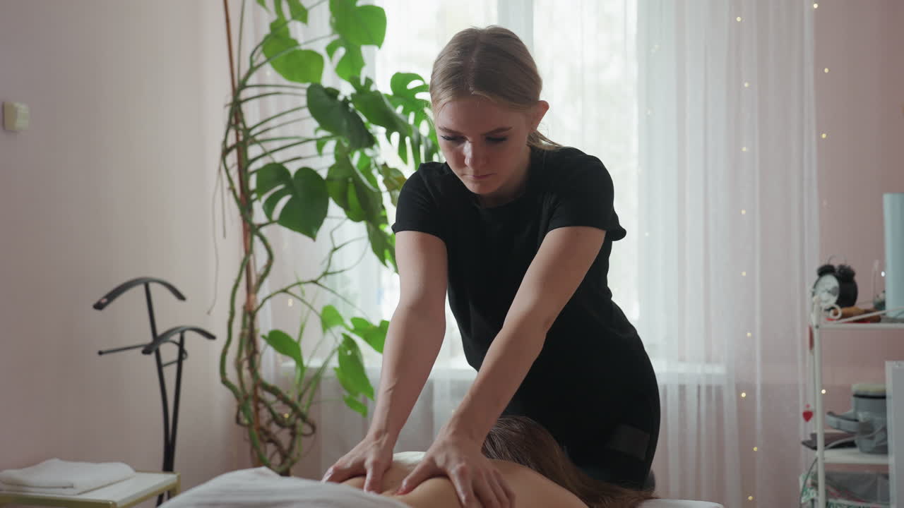 Massage therapist in black outfit gently pressing hands on client s back during therapeutic session in softly lit room with large green indoor plant and sheer white curtain enhancing tranquil ambiance