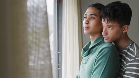 Happy-diverse-male-couple-embracing-and-smiling,-looking-through-window-in-living-room