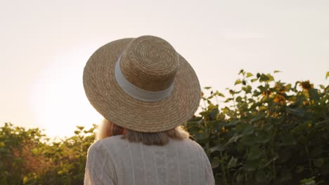 widok z tyłu pięknej młodej kobiety w słomkowym kapeluszu i białej sukience w stylu wiejskim, uśmiechającej się i tańczącej na polu słoneczników o zachodzie słońca