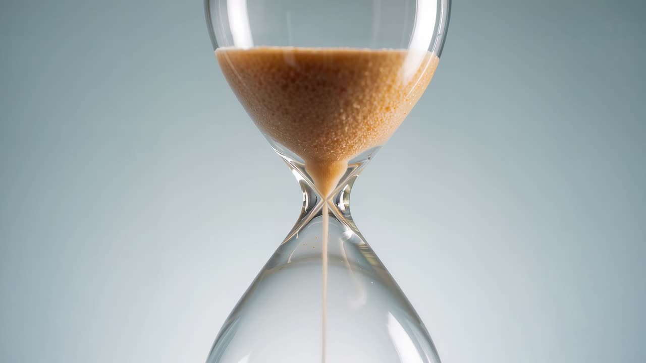 Gravity pulling tan sand trickling through glass hourglass against gray backdrop, marking time