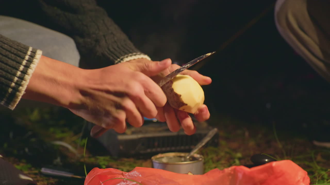 Close up of individual peeling Irish potato using knife during outdoor camping at night, hands working steadily over plastic bag with canned food nearby, surrounded by grass under warm light