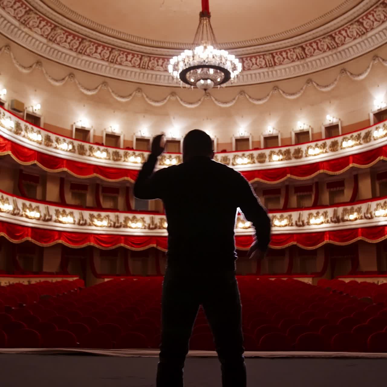 Back view of actor on stage. Man practicing speech with gestures in empty auditorium with red chairs. Performer is reheases in theater