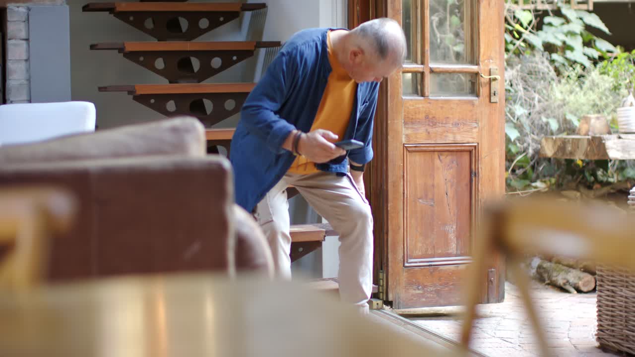 Anziano uomo asiatico che scende scale sospese, si ferma, si siede su una panca all'ingresso di casa e legge il telefono.