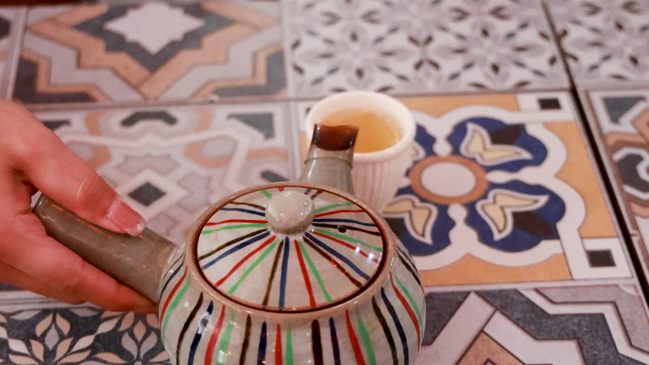 A hand pours tea from a striped teapot into a cup on a patterned tile surface
