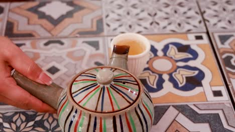 a hand pours tea from a striped teapot into a cup on a patterned tile surface