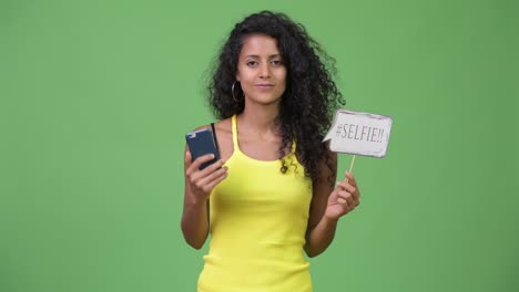 young beautiful hispanic woman holding phone and selfie paper sign