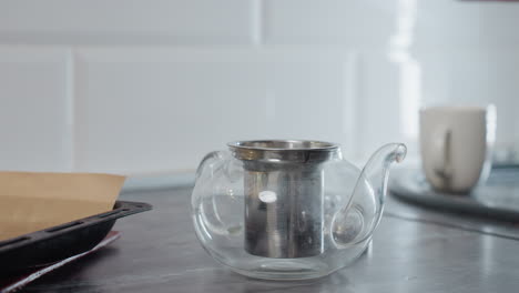 primer plano de una tetera de vidrio con filtro que se llena con sustancia de un paquete, alguna sustancia derramándose sobre la mesa, evocando una atmósfera de cocina hogareña